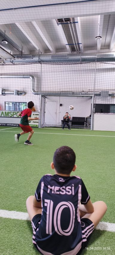 Junge sitzt auf einem Fußballfeld, schaut einem anderen Jungen beim Spielen zu.